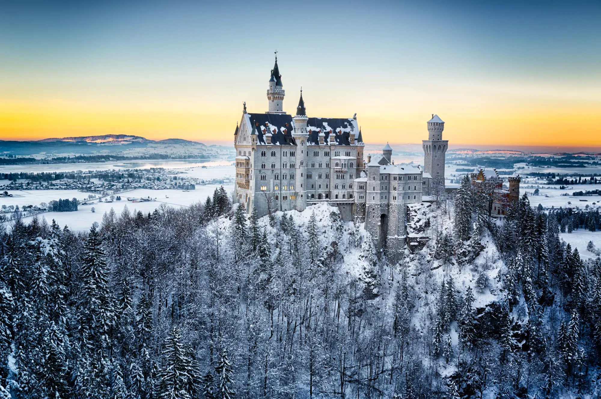 Wide angle shot Neuschwanstein Castle in the winter, showing its surrounding land covered in snow and a forest of fir trees in the forefront of the image. Behind the castle is a orange sky where the sun is setting