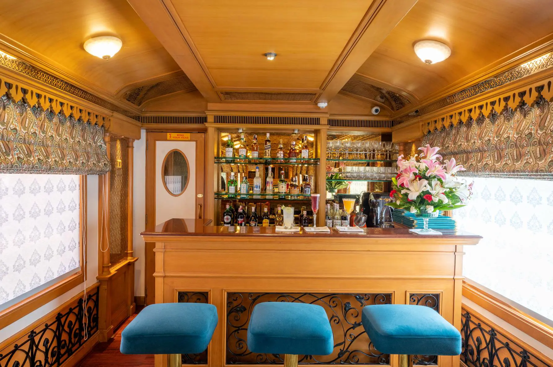 The Deccan Odyssey bar carriage with three teal bar stools, three drinks on the counter, and a range of bottles displayed behind the bar