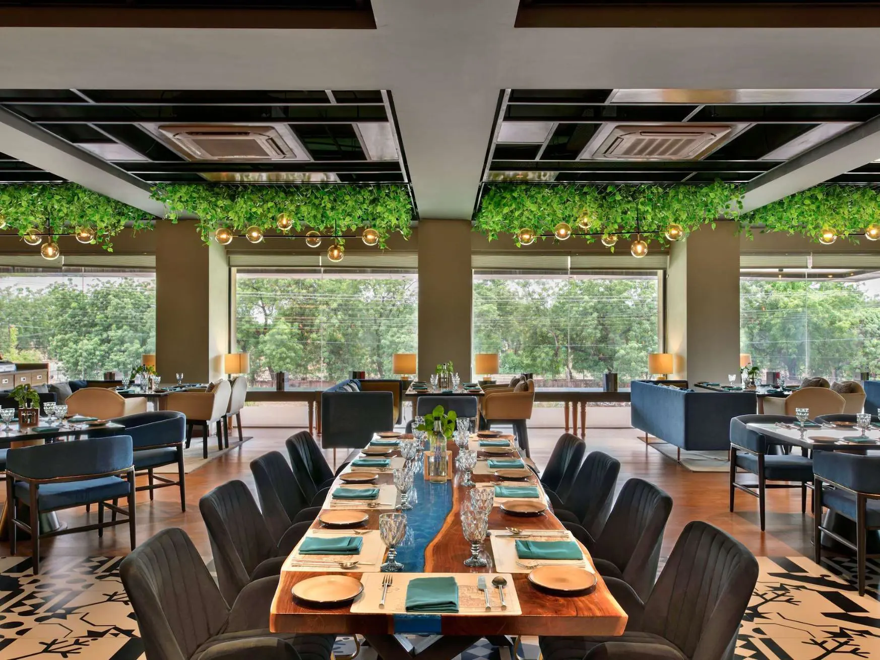 Modern restaurant at Novotel Jodhpur with large windows, hanging greenery, elegant table settings, and a mix of comfortable seating overlooking trees outside