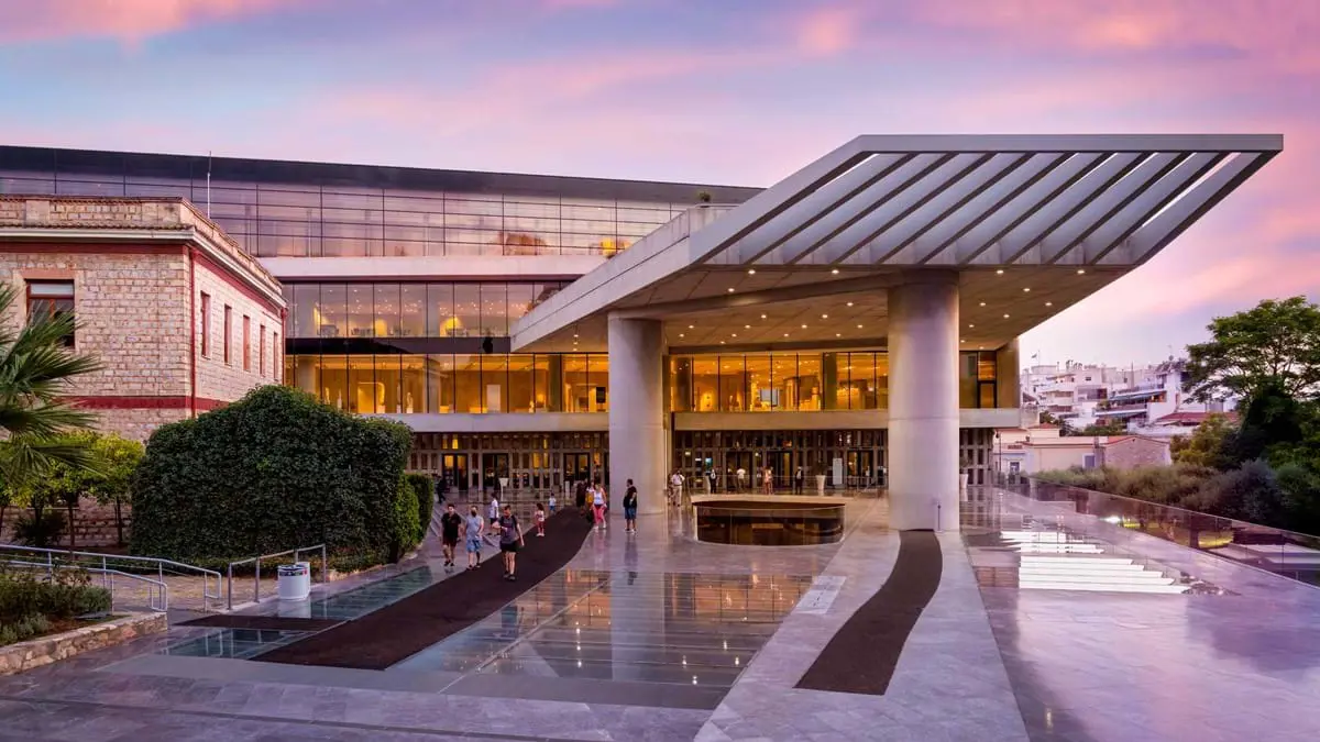 Modern building with marble flooring outside, and extended section on the right and windows. Purple sunset in the background