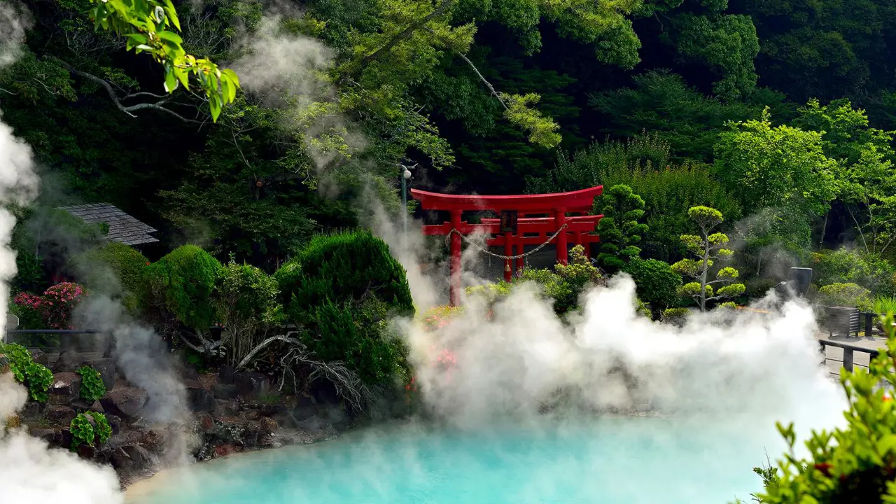 A steaming turquoise hot spring surrounded by lush greenery and a red torii gate in Beppu, Japan
