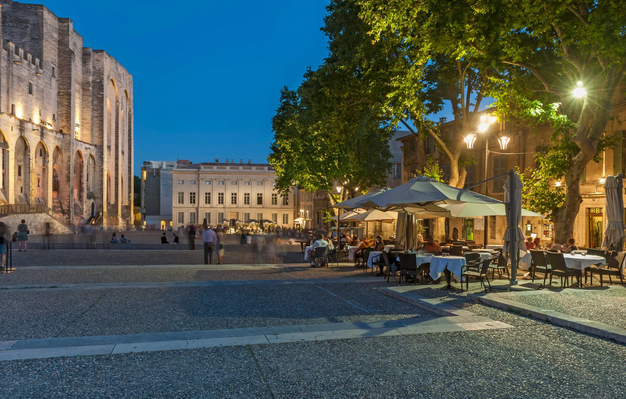 Outdoor seating outside The Palais Des Papes, that has white umbrellas, street lighting and trees above. A cream, wide manor is at the end of the road, and the edge of palace on the left.