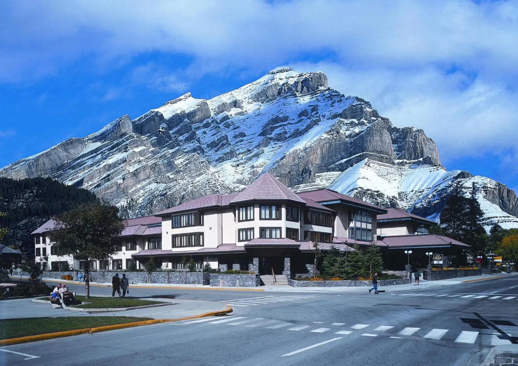 Elk + Avenue Hotel in Banff with a backdrop of rugged, snow-dusted mountains and a few pedestrians crossing the street