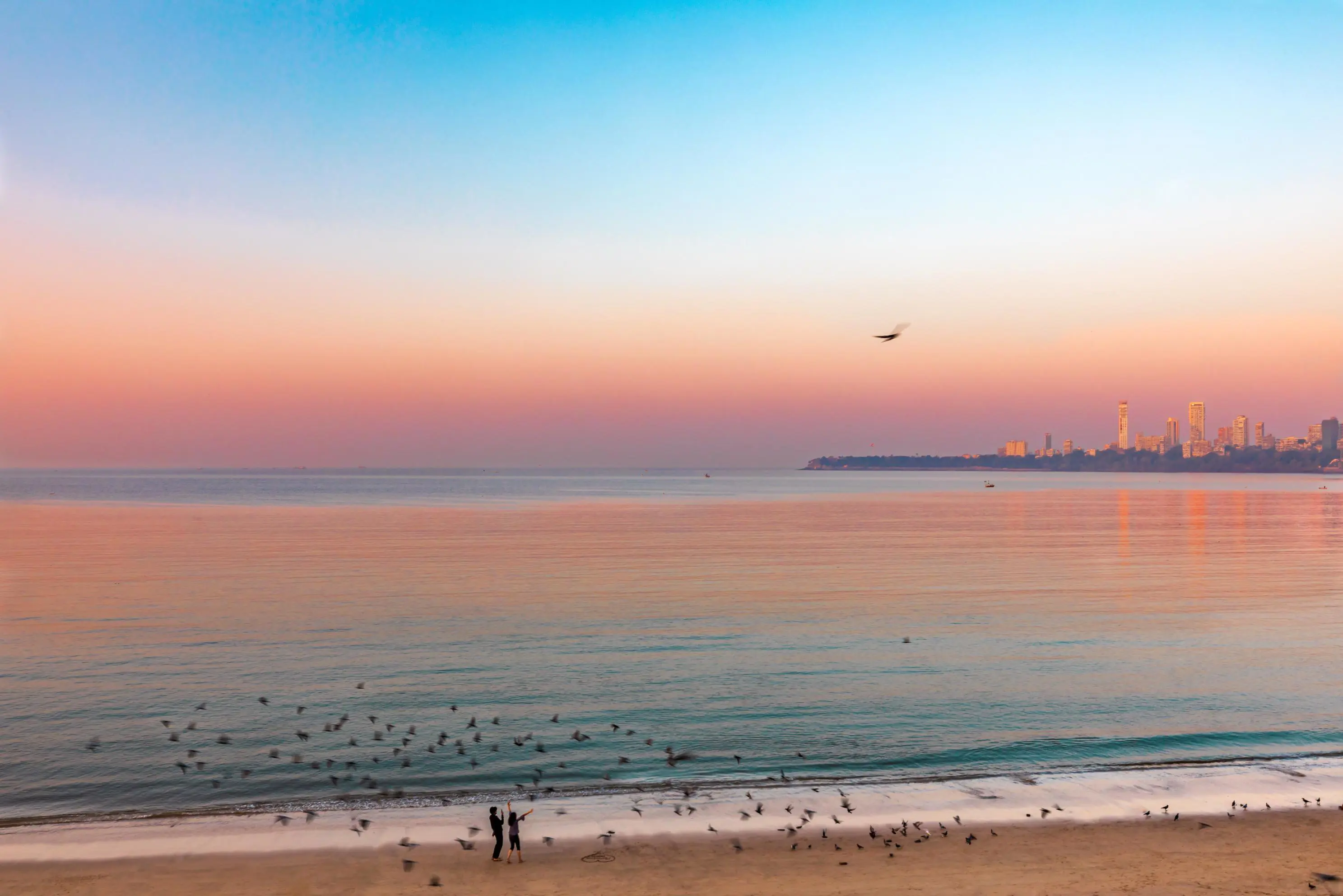 Mumbai seafront at sunset on a calm day, with birds flying over the water, two people standing on the beach, and the city skyline in the background