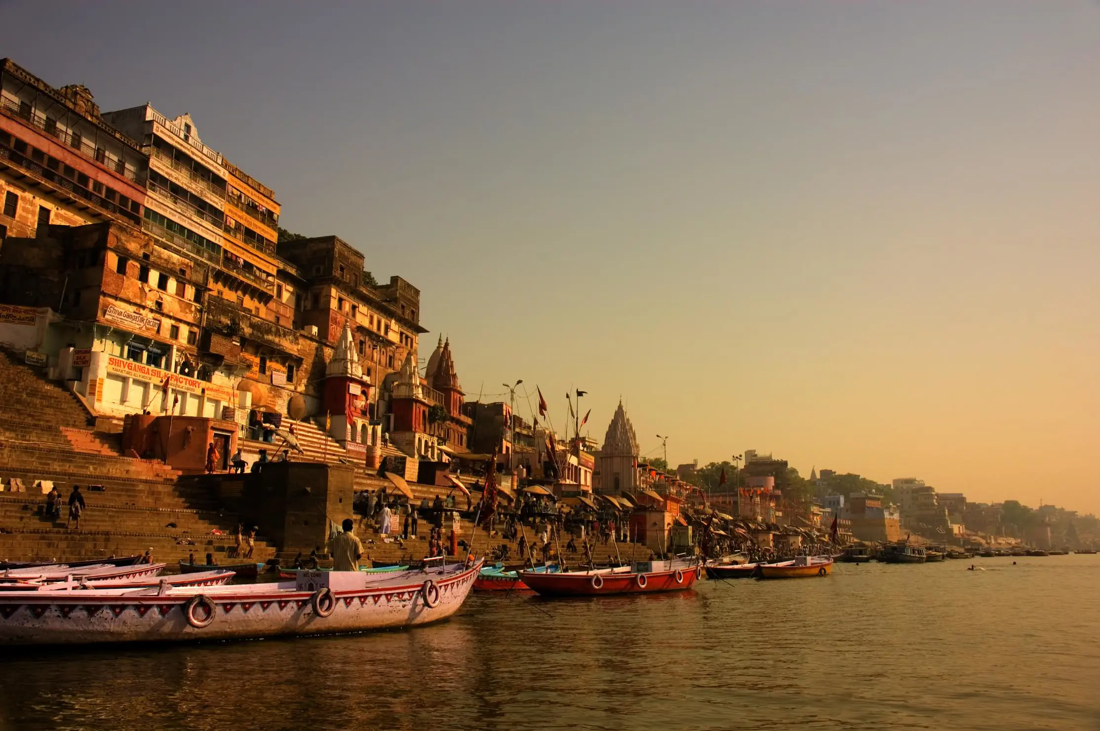 Boats anchored along the ghats of Varanasi at sunrise, with ancient sandstone buildings and temples glowing in golden light by the River Ganges