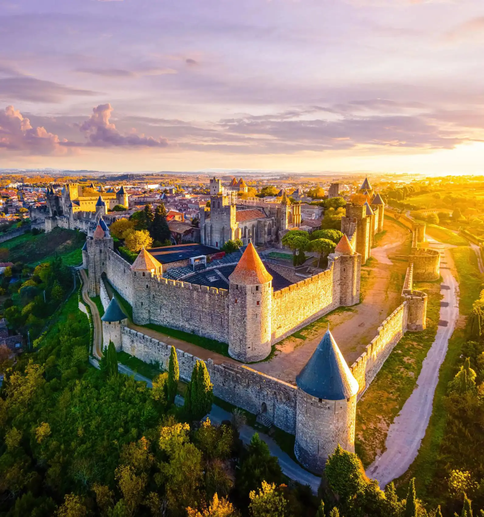 Bird's eye view of the Castle of Carcassonne at sunset, surrounded by trees and grass, and a pathway that loops around the castle. The castle has towers with red and blue turrets, and a distant view of a town can be seen behind it
