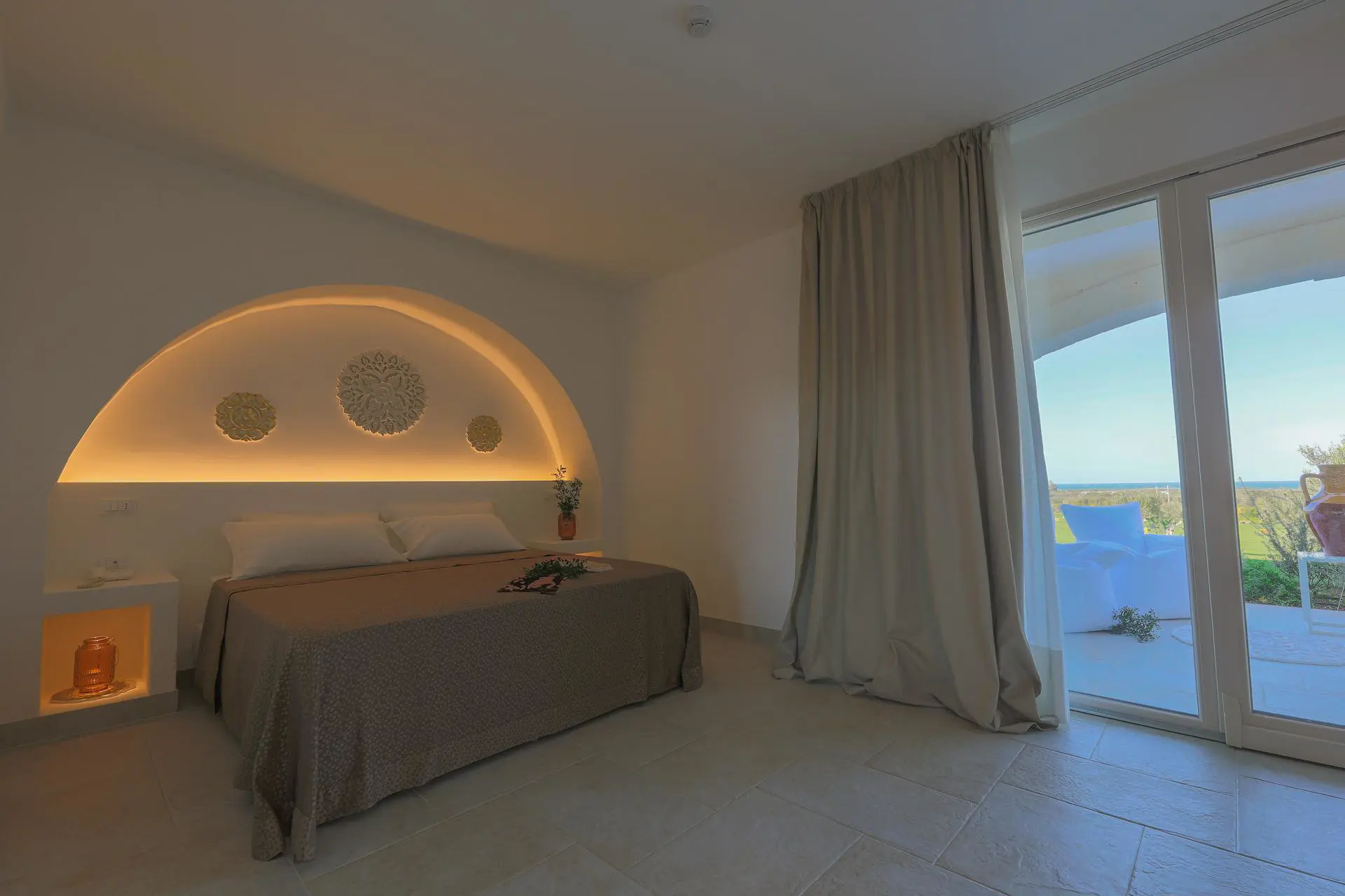 Modern bedroom at Masseria Casseli Hotel, Italy, with warm lighting, arched headboard, and view of the garden and sea through large glass doors