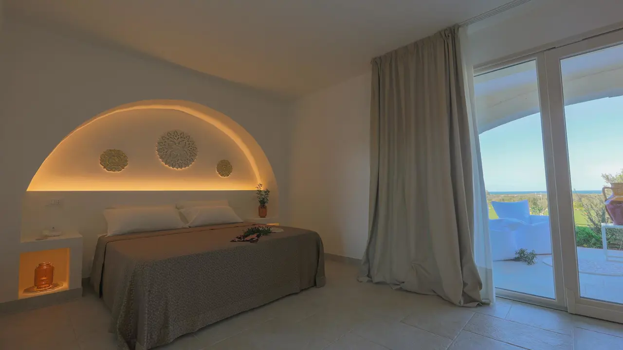 Modern bedroom at Masseria Casseli Hotel, Italy, with warm lighting, arched headboard, and view of the garden and sea through large glass doors