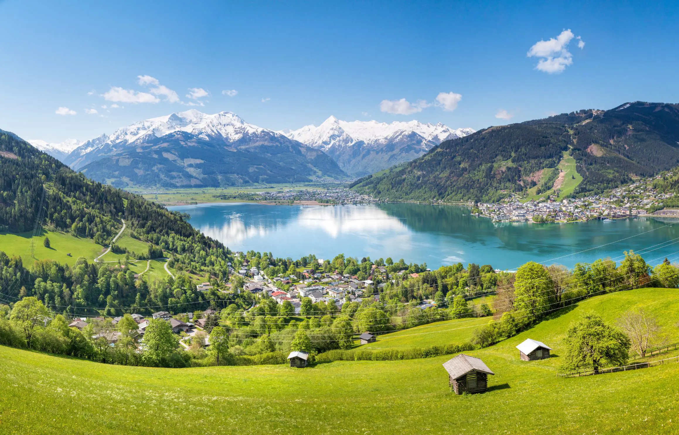 View of a large lake from a mountain, with houses and trees at the bottom. Another town can be seen on the other side of the lake to the right and centre of the image, and mountains with some snow on the top can be seen in the distance.