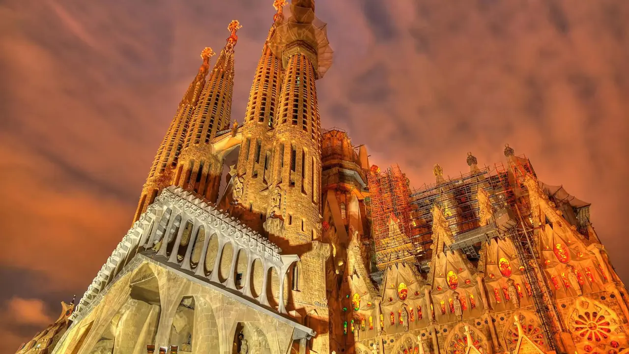 Night view of the illuminated Sagrada Família basilica in Barcelona, Spain, showcasing its intricate architecture and towering spires against a dramatic, cloudy sky