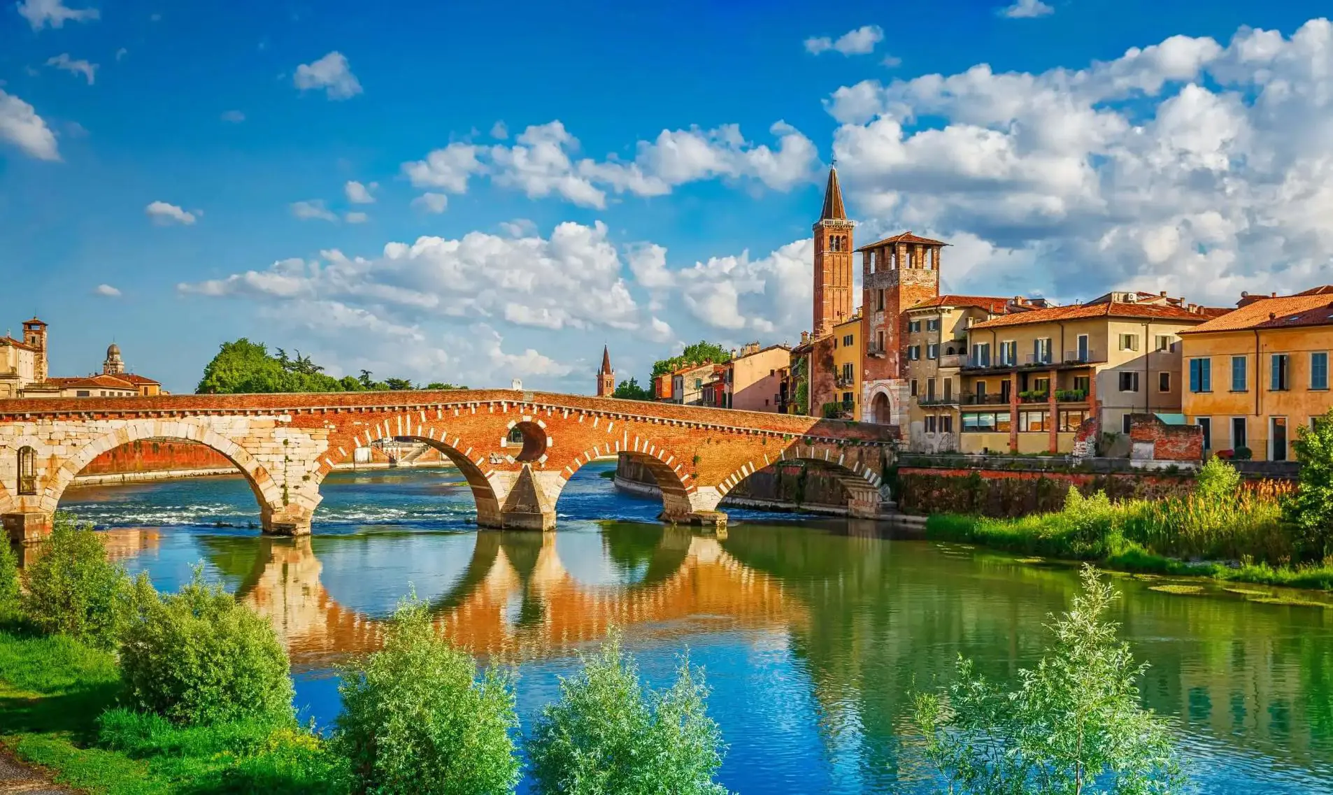 Scenic view of the historic Ponte Pietra bridge crossing the Adige River in Verona, Italy, with colourful old buildings and church towers under a bright blue sky with scattered clouds