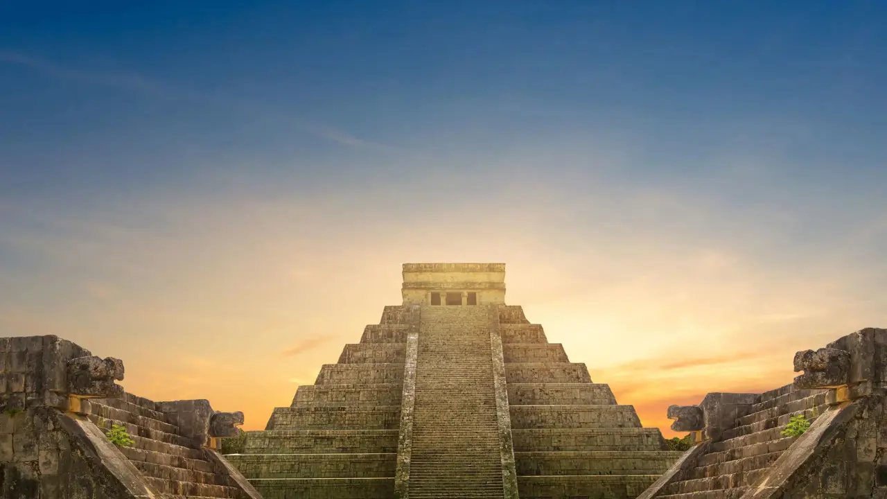 The ancient Mayan pyramid of Chichen Itza at sunset, with stairs leading up the centre and a clear sky fading from orange to deep blue