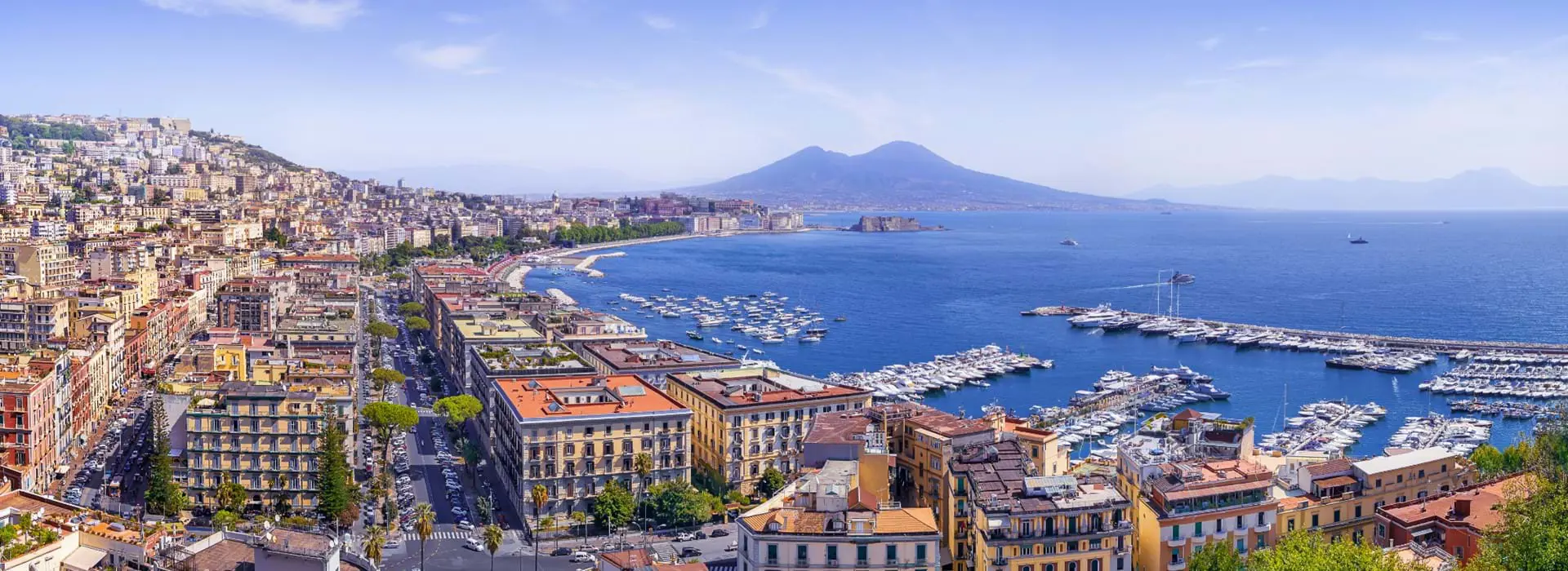The Bay of Naples with Mount Vesuvius in the distance, lined with waterfront buildings and apartments, and a marina filled with boats