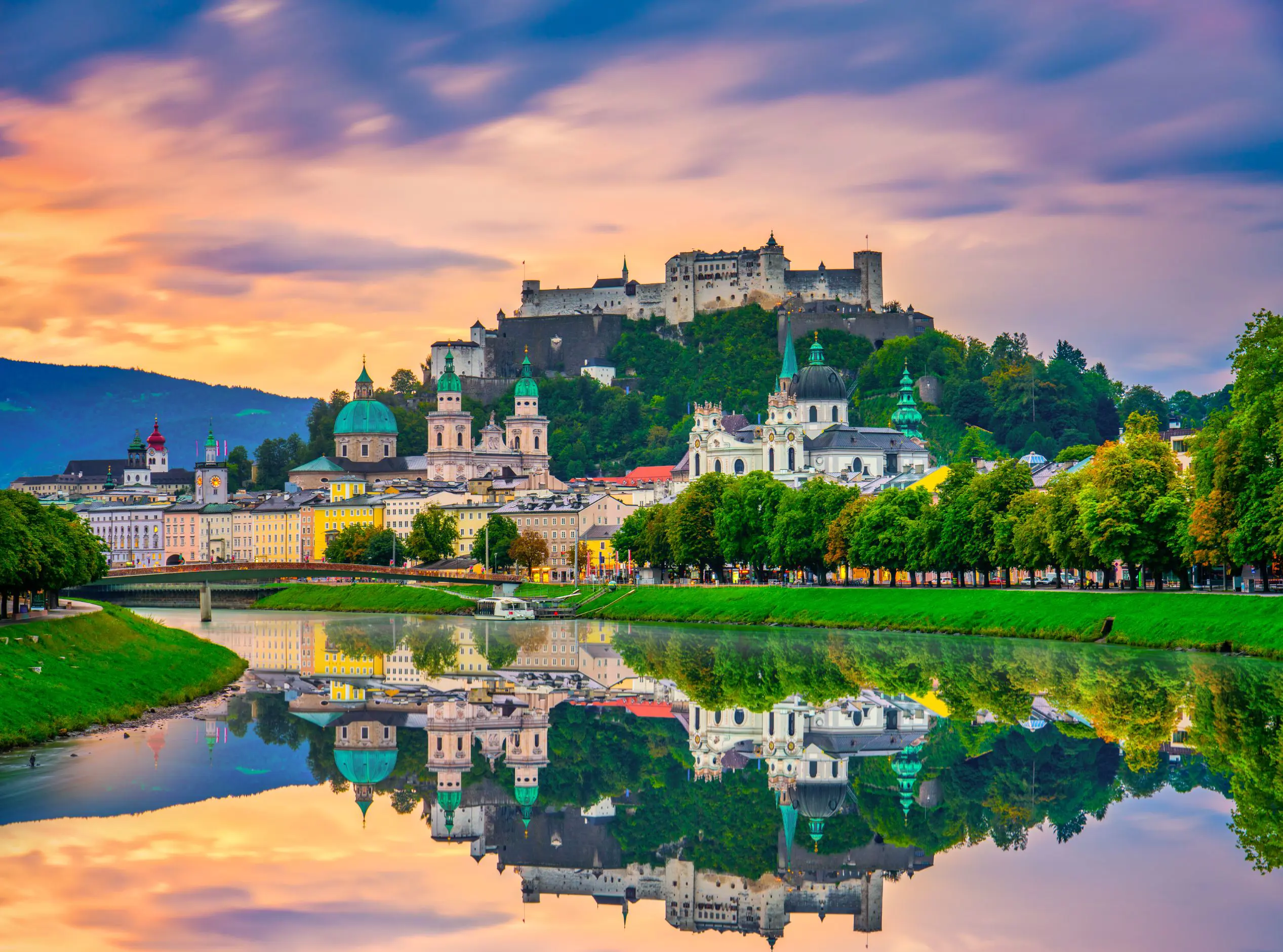 View of Salzburg from the river, showing houses along the river bed, churches and historical landmarks with turquoise turrets behind them, and the fortress on the top of the hill. There is a clear reflection of the city in the river at the forefront of the image, and the sky is a purple and orange sunset