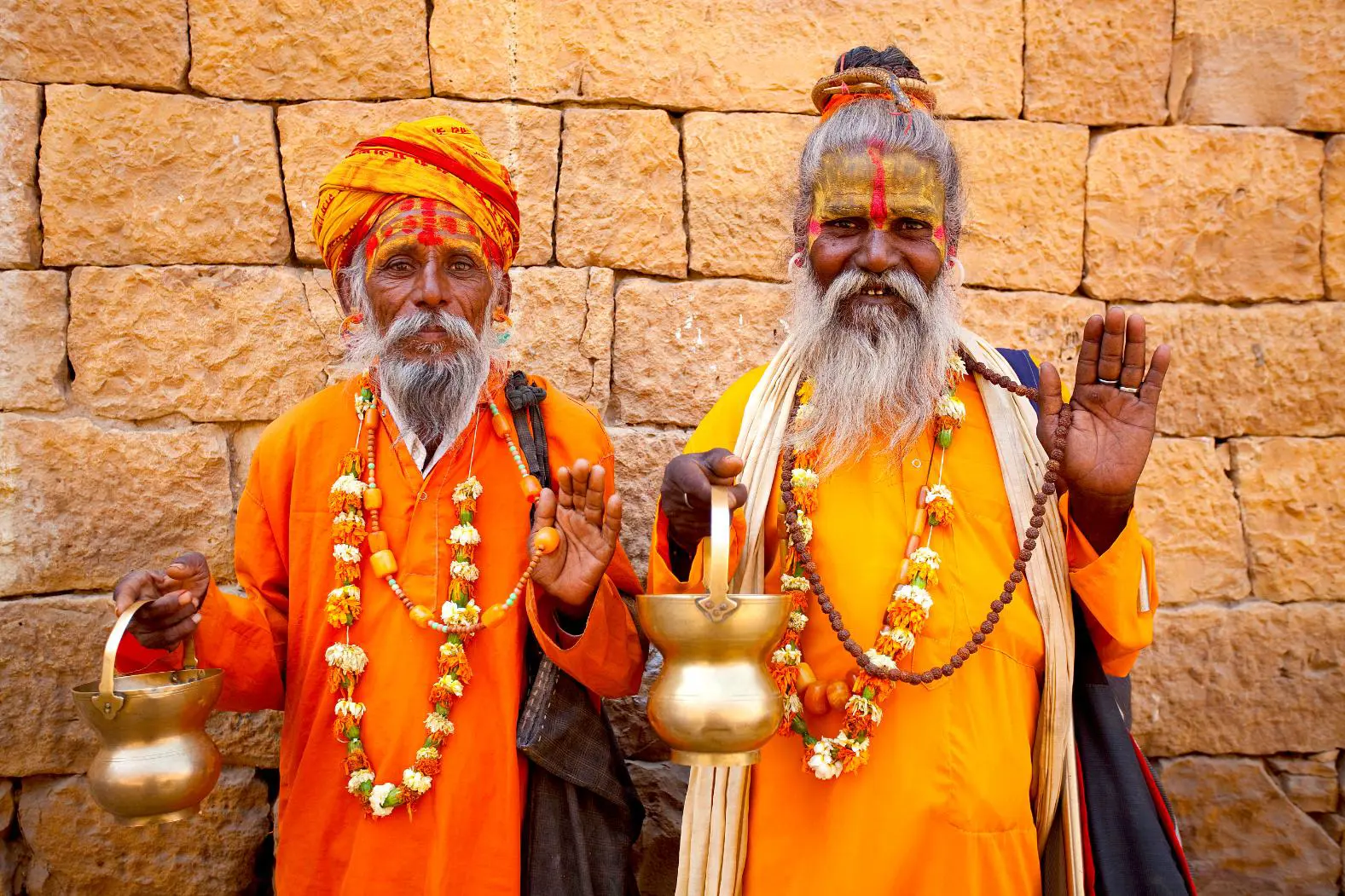 Two elderly Jain priests in orange robes and garlands stand before a sandstone wall, holding brass pots in Rajasthan in India