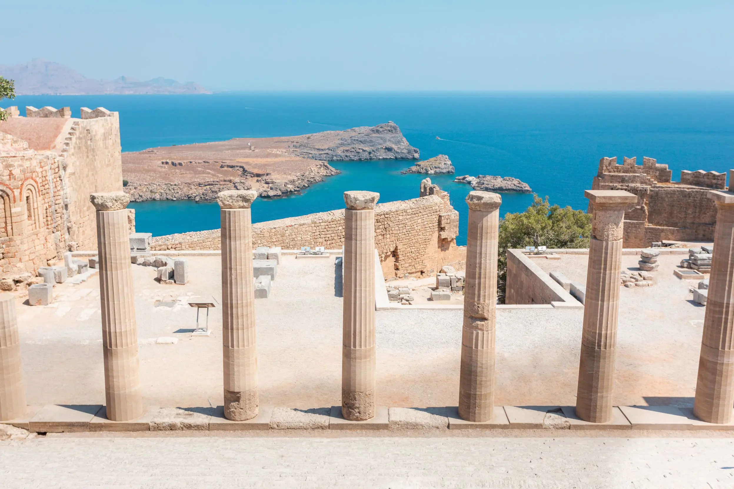 Stone pillars lined up in the forefront, with the ruins of Lindos city in front, and the view of the sea.
