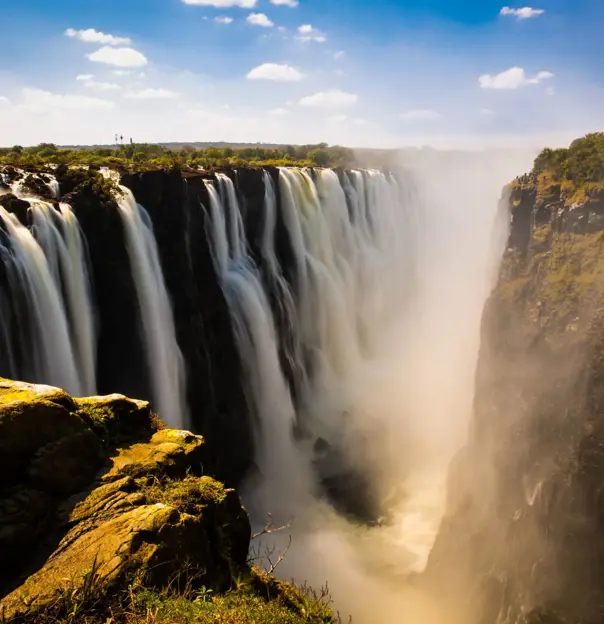 Victoria Falls cascading powerfully over cliffs, surrounded by lush greenery under a bright blue sky with scattered clouds