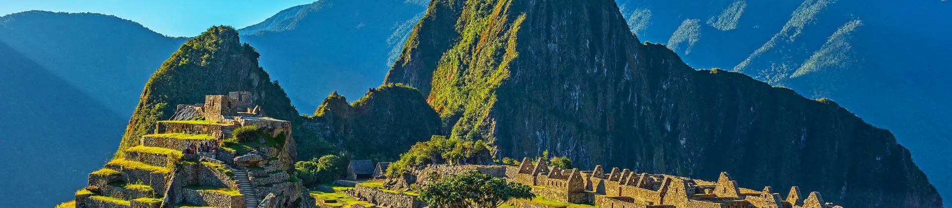 A panoramic view of Machu Picchu in Peru, showing ancient Incan ruins built into a green mountain ridge under a clear blue sky, with Huayna Picchu towering in the background