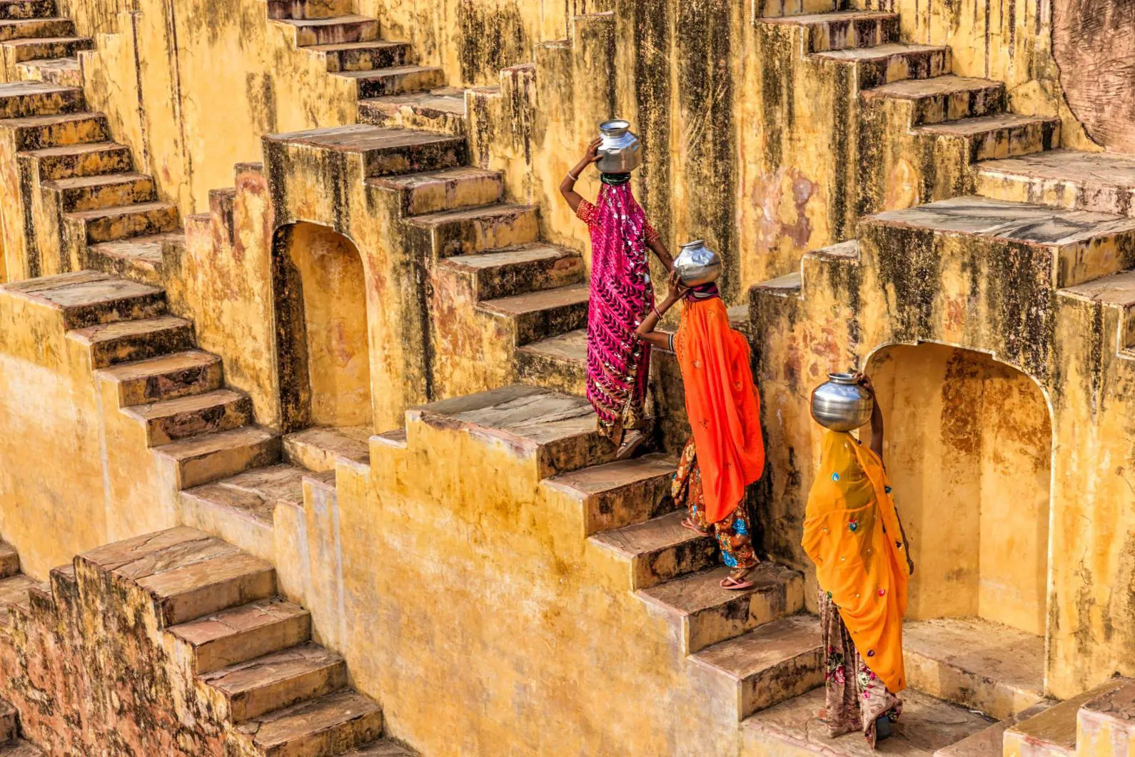 Three women in traditional Rajasthani dress walking up the stone steps at Chand Baori, each carrying a metal water container on her head