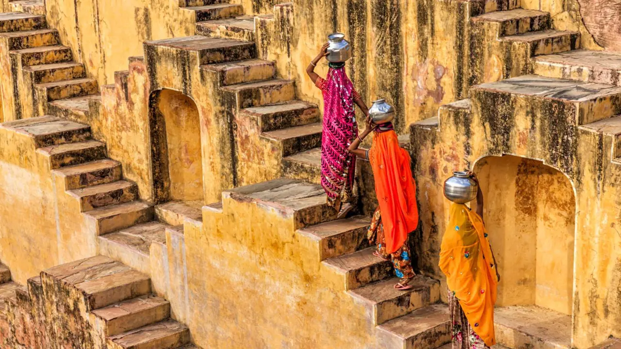 Three women in traditional Rajasthani dress walking up the stone steps at Chand Baori, each carrying a metal water container on her head