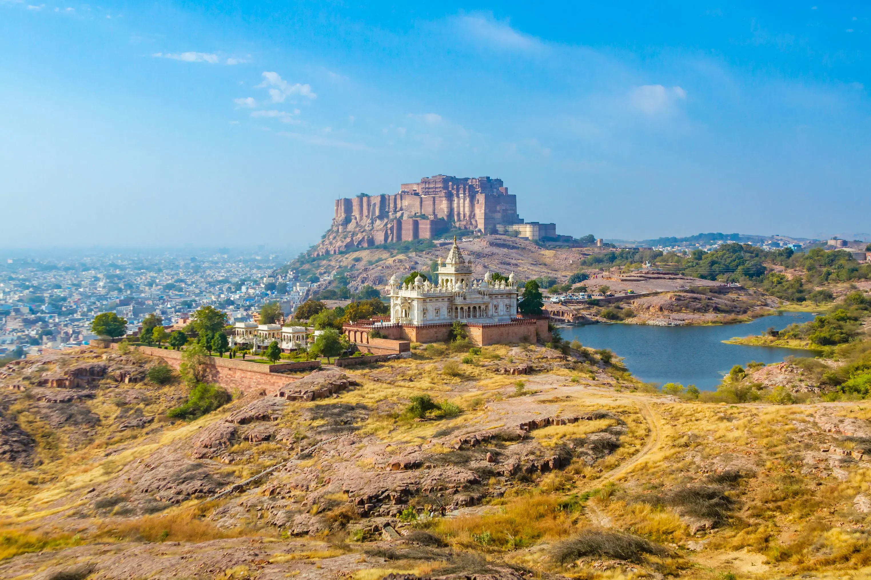View of the white marble Jaswant Thada memorial and Mehrangarh Fort rising above Jodhpur, with a lake and cityscape in the background under a clear blue sky