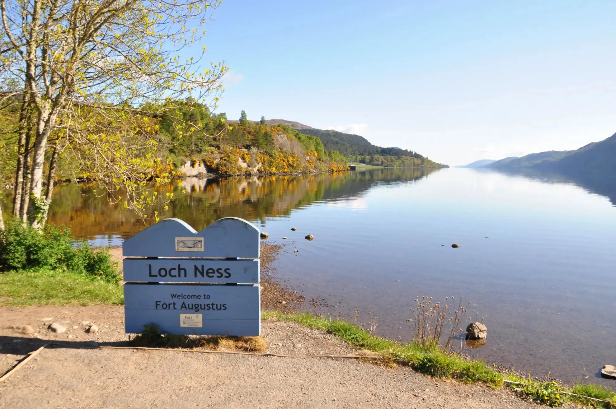 Calm waters of Loch Ness reflecting the surrounding hills under a clear blue sky, with a sign welcoming visitors to Fort Augustus on the shore
