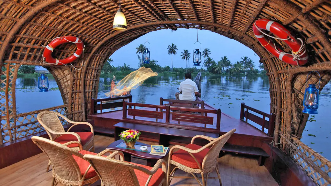 A cosy houseboat with wicker furniture sails through calm backwaters as a man casts a fishing net under palm trees at dusk