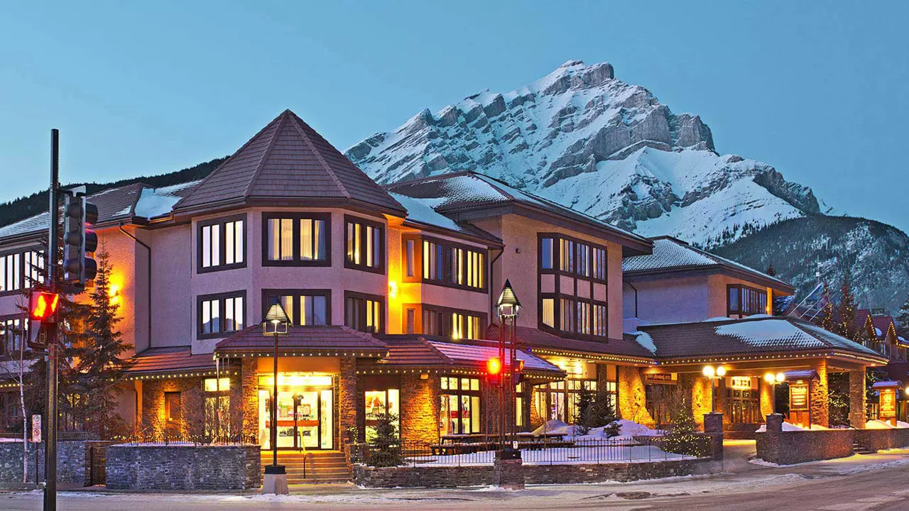 The exterior of Elk & Avenue Hotel in Banff at night, with warmly lit windows and snow-capped mountain in the background