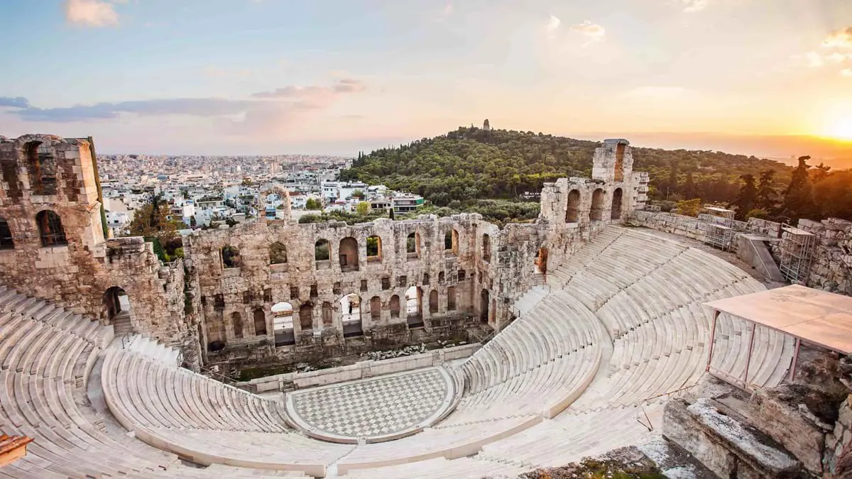 High angle view of the ruins of an amphitheatre, with the view of a city in the distance and forested land to the left.
