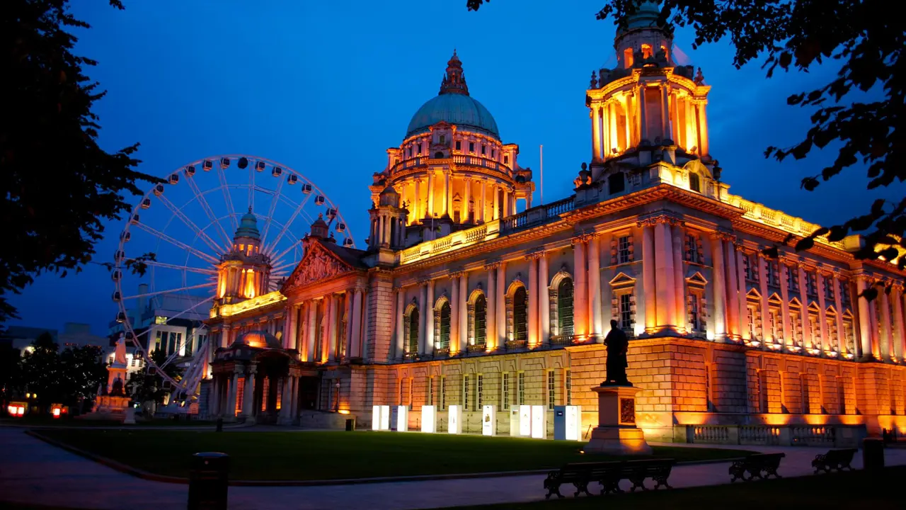Belfast City Hall illuminated in warm lights at dusk, with a large ferris wheel behind it against a deep blue sky