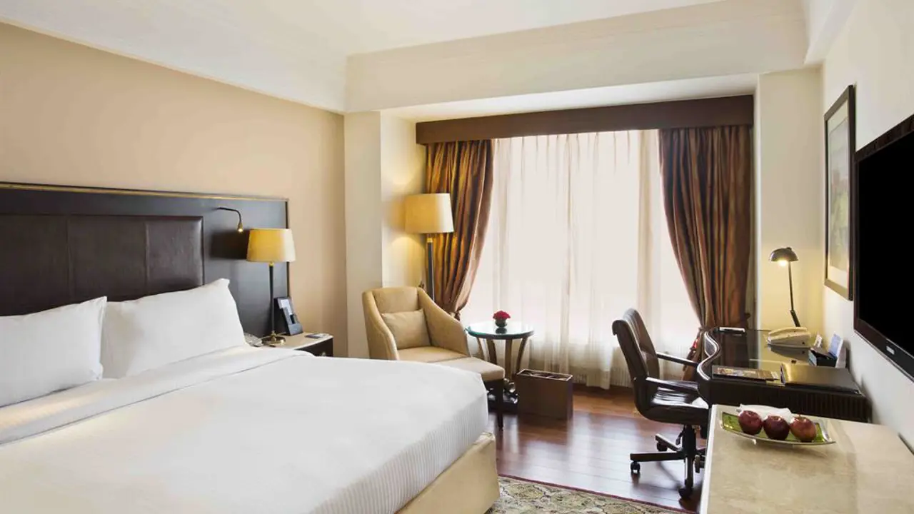 Modern double room at Eros Hotel in New Delhi with a large bed, work desk, armchair and floor-to-ceiling curtains letting in natural light