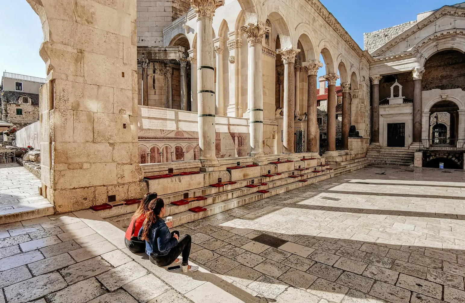 Two women sit on the iconic steps of the Peristyle courtyard in Diocletian’s Palace, surrounded by ancient Roman columns and white stone architecture