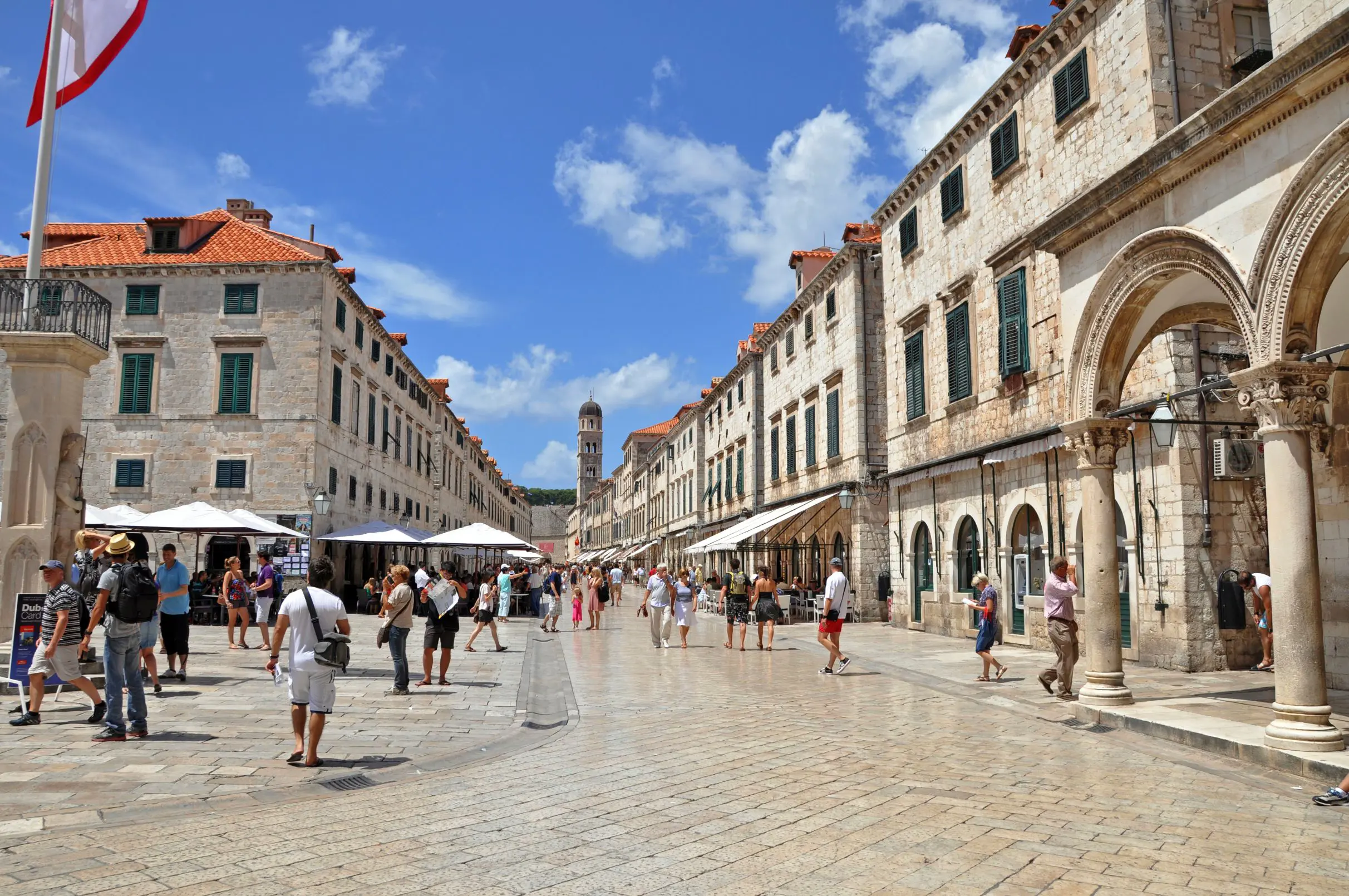 View of a street in the Old Town, Dubrovnik, bricked buildings with orange roofs either side and people in the street. White umbrellas all down the road an a tower at the end. Blue sky with some clouds.
