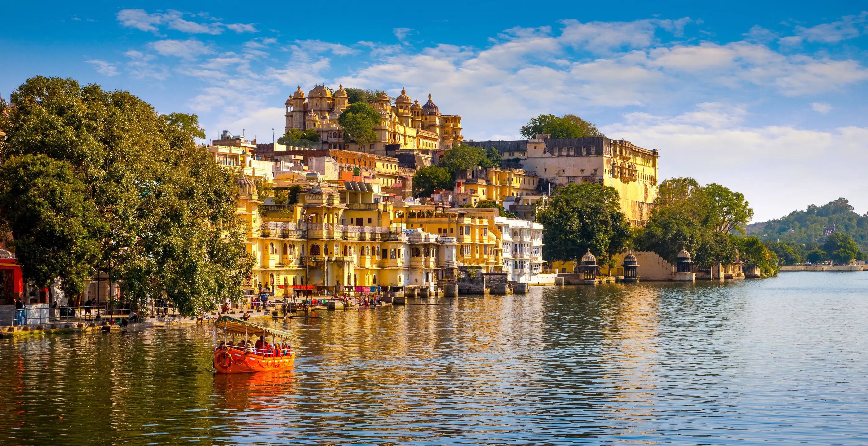 Scenic view of Udaipur’s City Palace and colourful lakeside buildings reflected in Lake Pichola, with a small red boat on the water under a bright blue sky