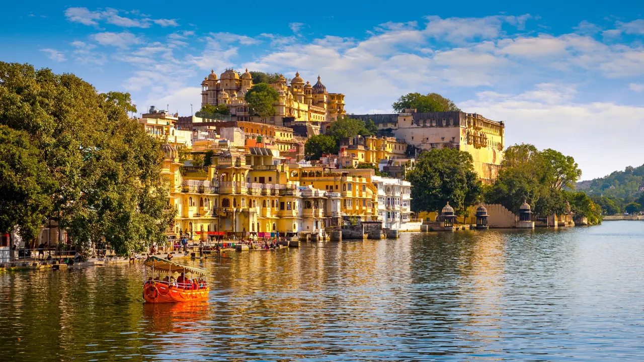 Scenic view of Udaipur’s City Palace and colourful lakeside buildings reflected in Lake Pichola, with a small red boat on the water under a bright blue sky