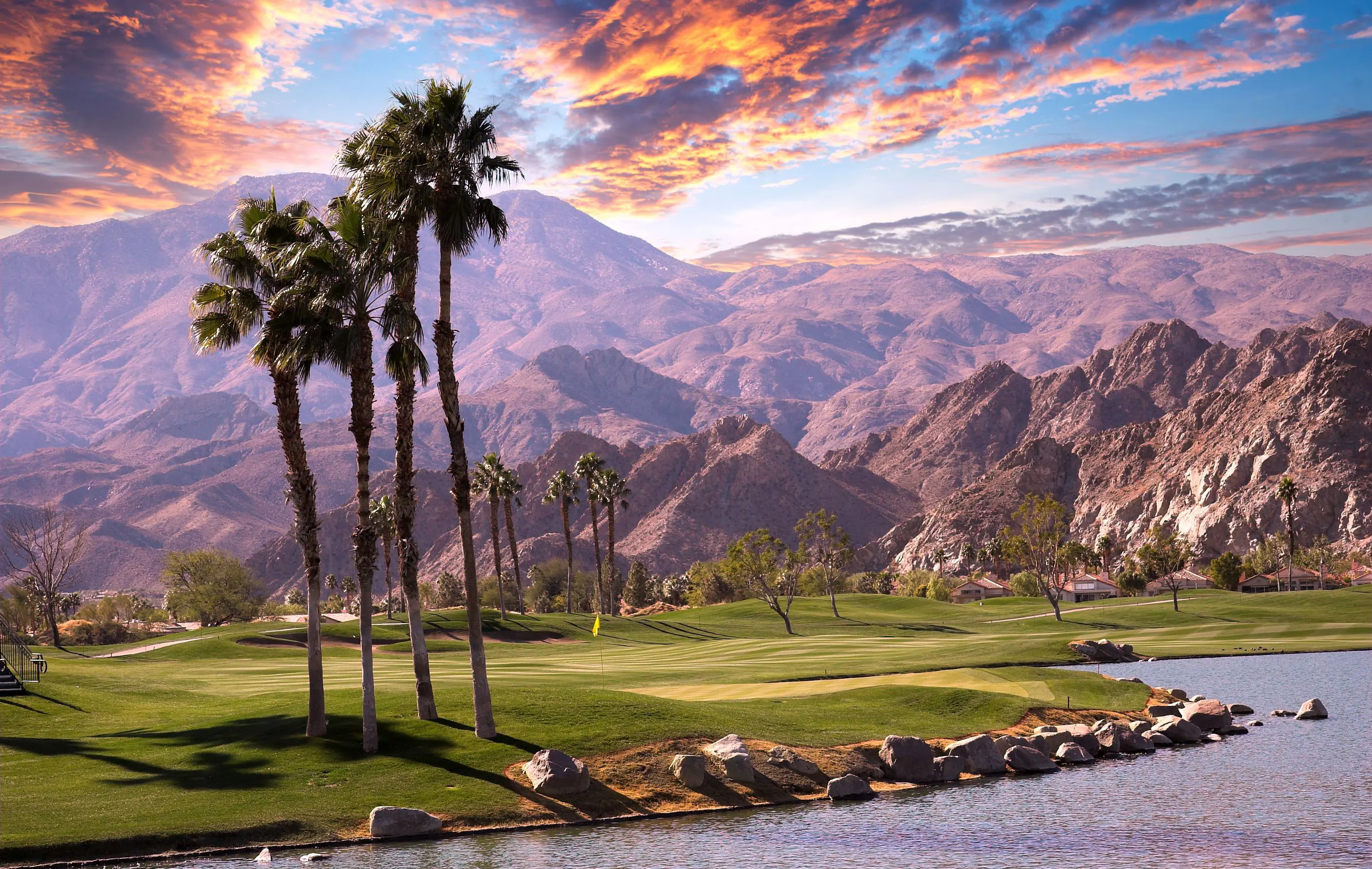 A scenic view of Palm Springs with mountains in the background and tall palm trees next to a small lake in the foreground