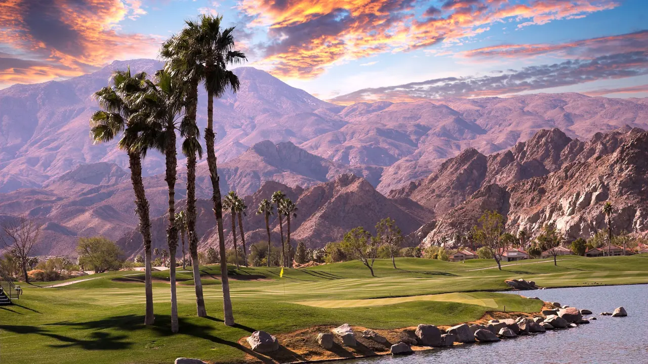 A scenic view of Palm Springs with mountains in the background and tall palm trees next to a small lake in the foreground