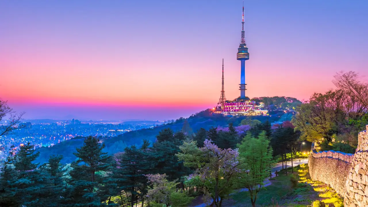 A view of Seoul Tower in South Korea at sunset, with warm orange and pink hues lighting up the sky and the cityscape below