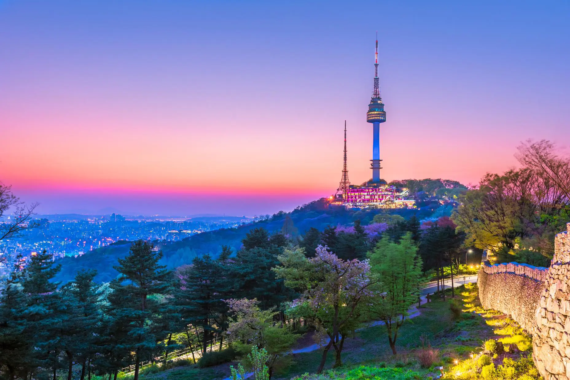 A view of Seoul Tower in South Korea at sunset, with warm orange and pink hues lighting up the sky and the cityscape below
