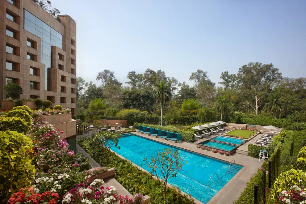 A view of the swimming pool and exterior of the Sheraton New Delhi, surrounded by landscaped gardens on a bright day