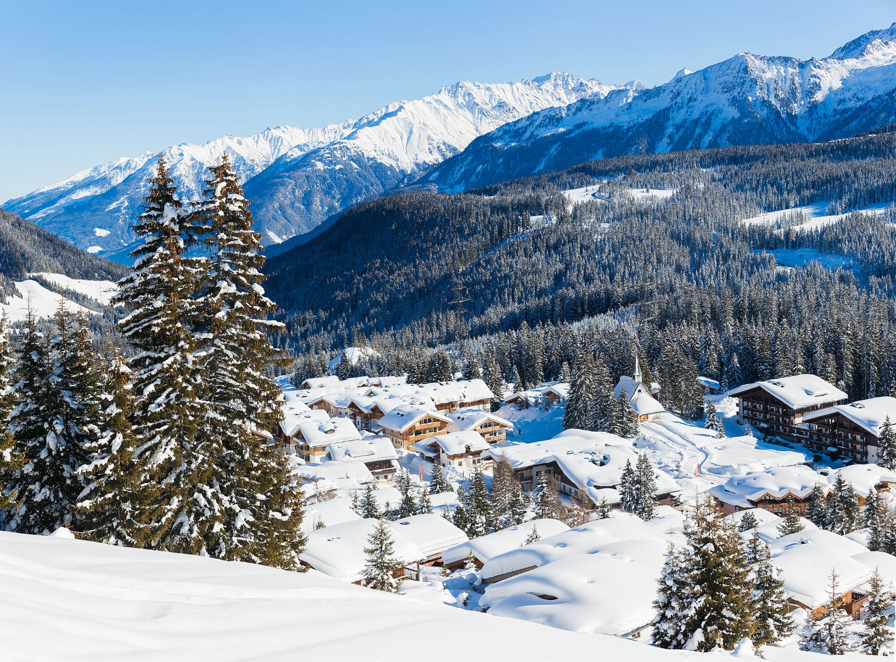 View of a village in the mountains in thick snow. With large fir trees in the left forefront, and smaller ones dotted around the village at the bottom the mountain. Above this a large forest of trees can be seen, going up the mountain, in front of large mountains covered in snow, in front of a light blue sky.