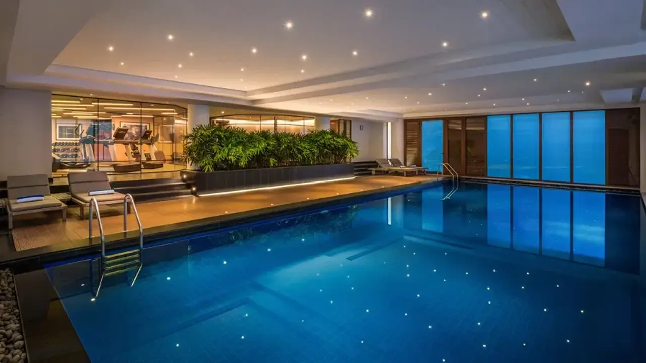 Indoor pool at The Residency Towers hotel in Pondicherry, illuminated by underwater spotlights, with sunbeds along the side and a glass window offering a view into the gym