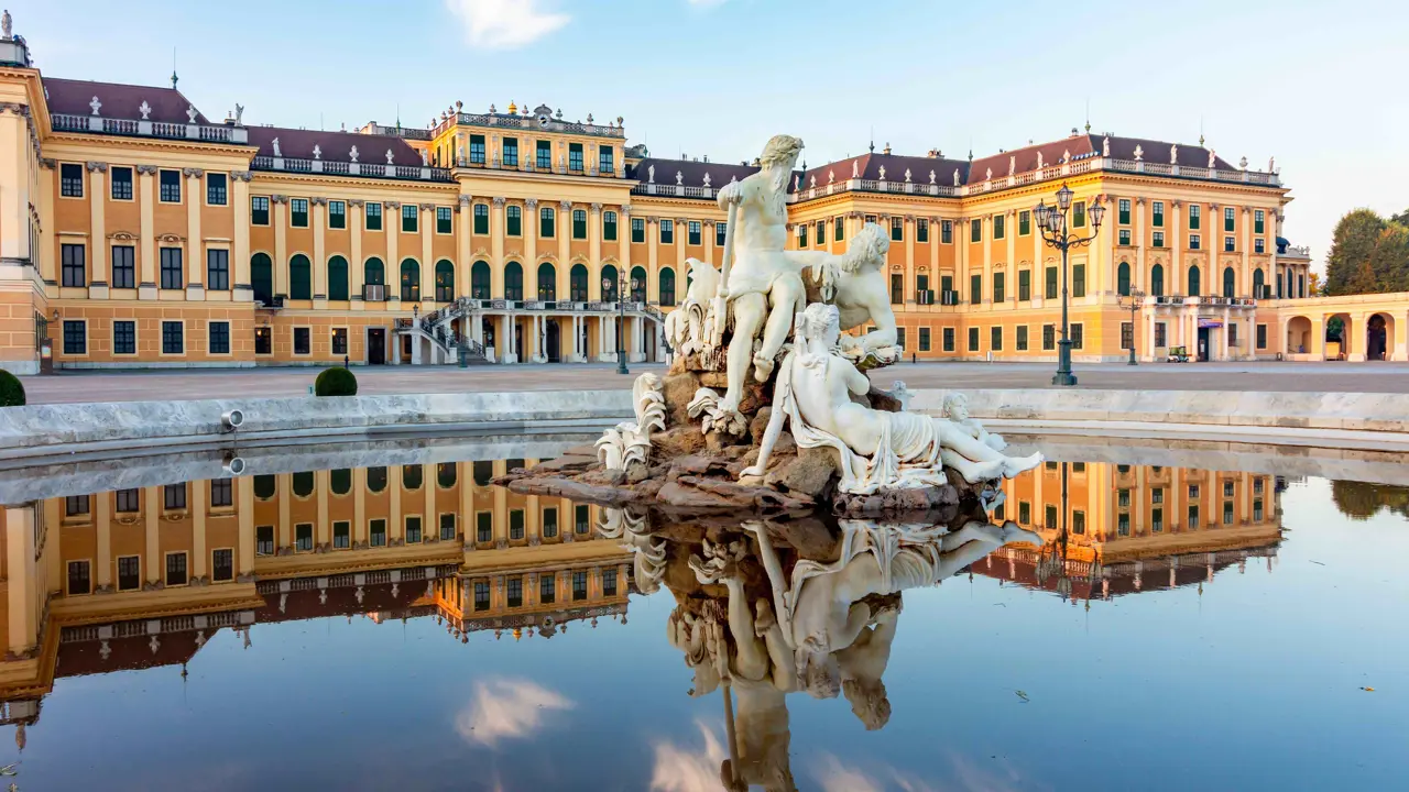 A fountain with a white statue of three people sitting on a rock, on top of water, where its reflection can be seen. Behind it is Schönbrunn Palace, yellow and beige wide building, in front of a blue sky with small clouds.