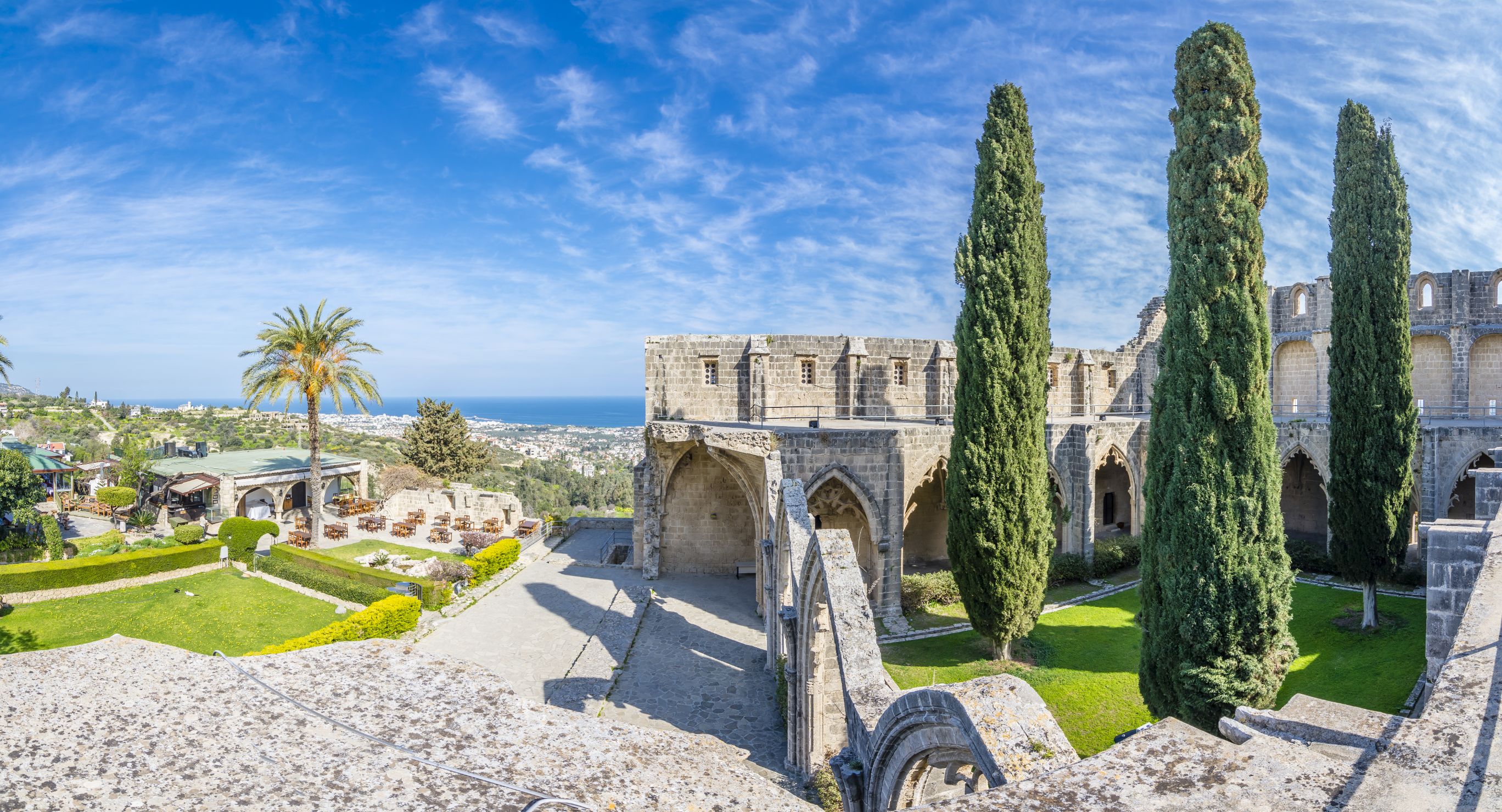 Stone ruins to the right, with a grassy area in the middle with three tall slim trees. Opposite, to the left is more grassy area with a palm tree and bushes. Behind this is a distant view of a town and the ocean, and a blue sky above.