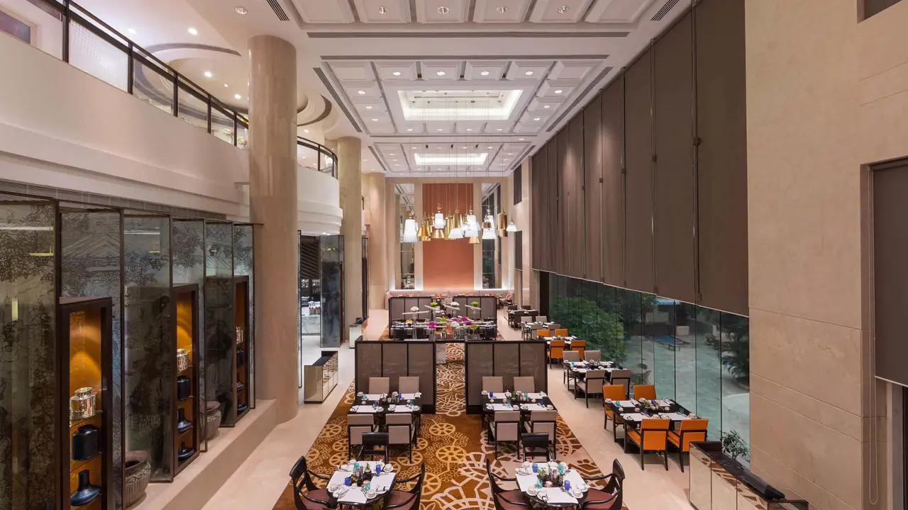 Modern dining area at Sheraton New Delhi, featuring high ceilings, elegant lighting, large windows, and neatly arranged tables in a spacious setting