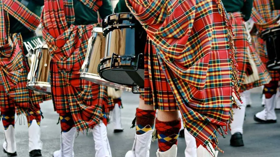 Close up of the bottom half of Scottish drum players in a parade wearing kilts, tartan shawls, kilt socks and white boots,holding drums 