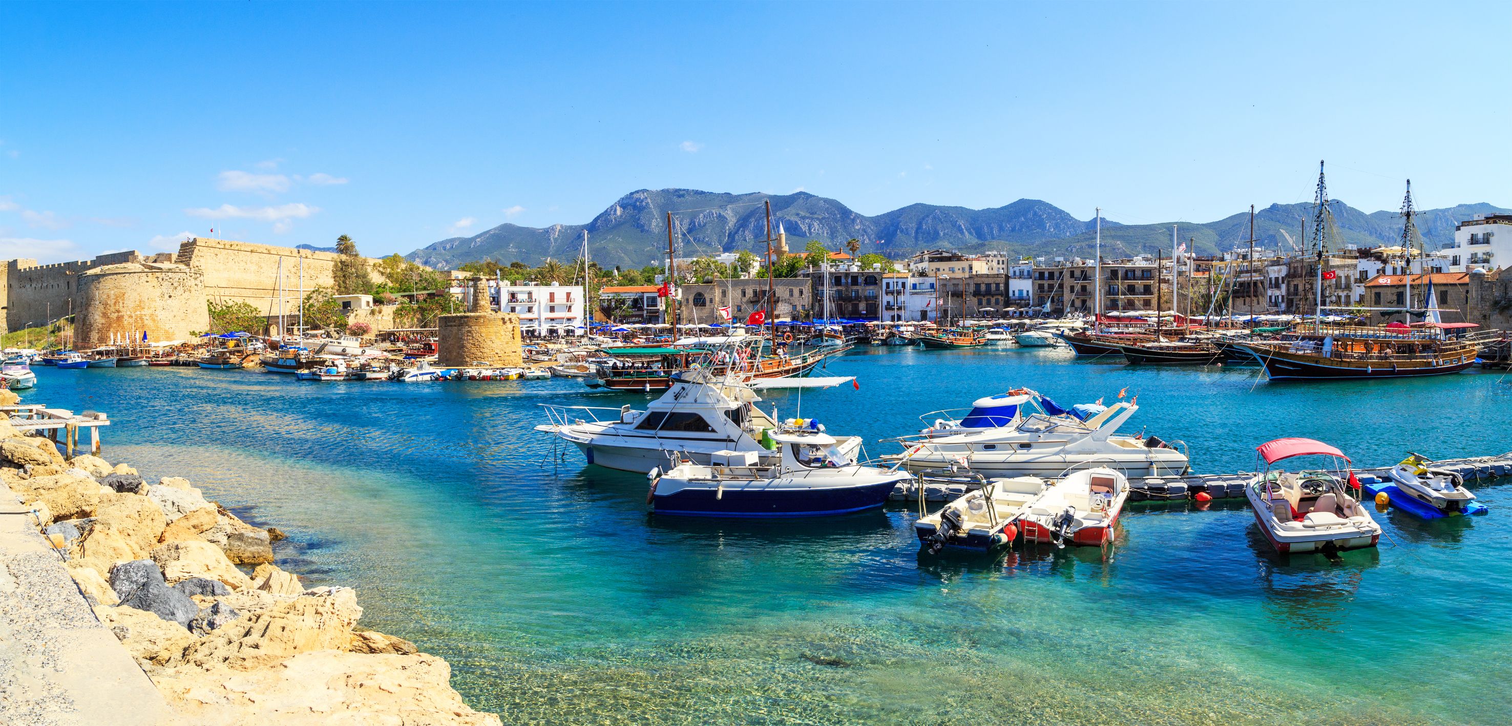 Boats docked at a harbour with bright blue water, buildings on the other side, and ruins of a castle to the left. Mountains in the distance in front of a blue sky.