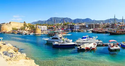 Boats docked at a harbour with bright blue water, buildings on the other side, and ruins of a castle to the left. Mountains in the distance in front of a blue sky.