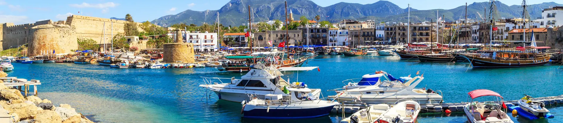 Boats docked at a harbour with bright blue water, buildings on the other side, and ruins of a castle to the left. Mountains in the distance in front of a blue sky.