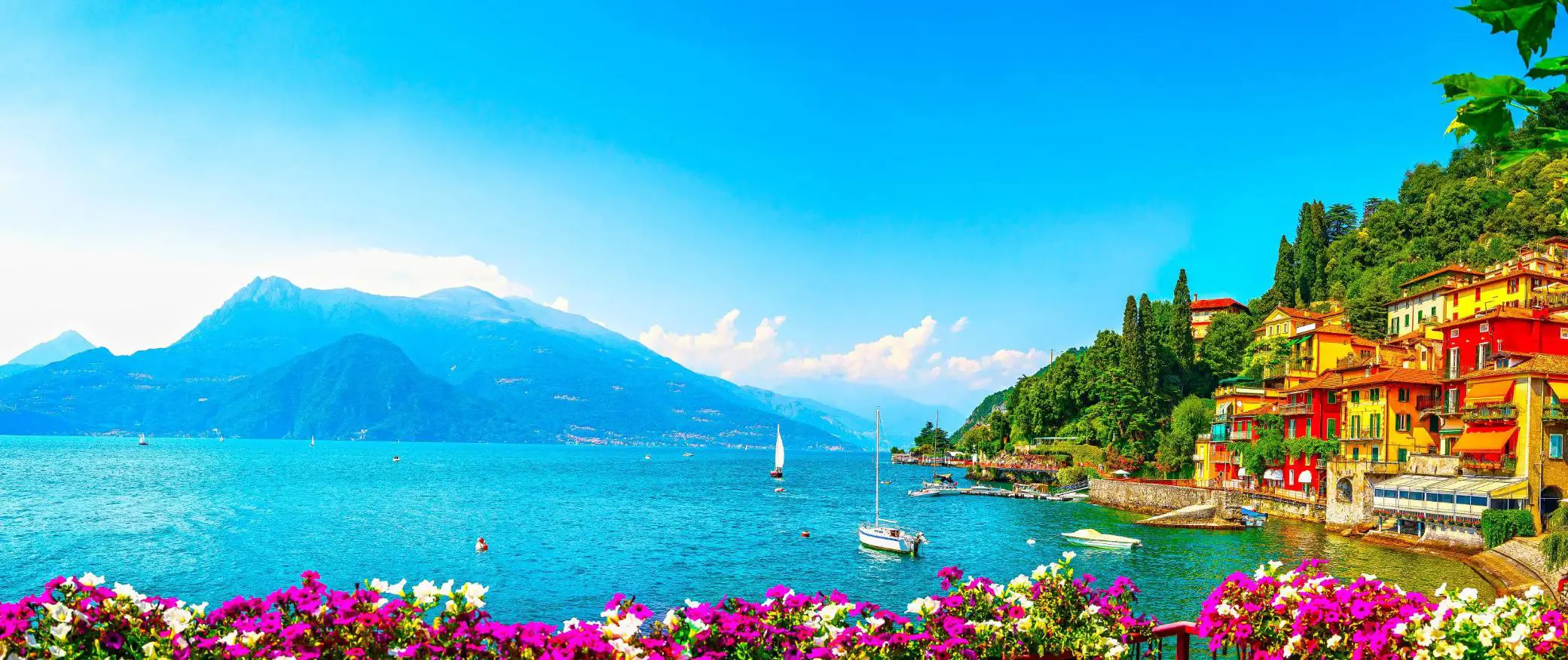Colourful hillside houses in Lake Como overlooking the lake, where a couple of boats gently float, with mountains in the background